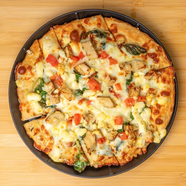 A sliced pizza with chicken, tomatoes, and cheese on a black plate, on a wooden surface.