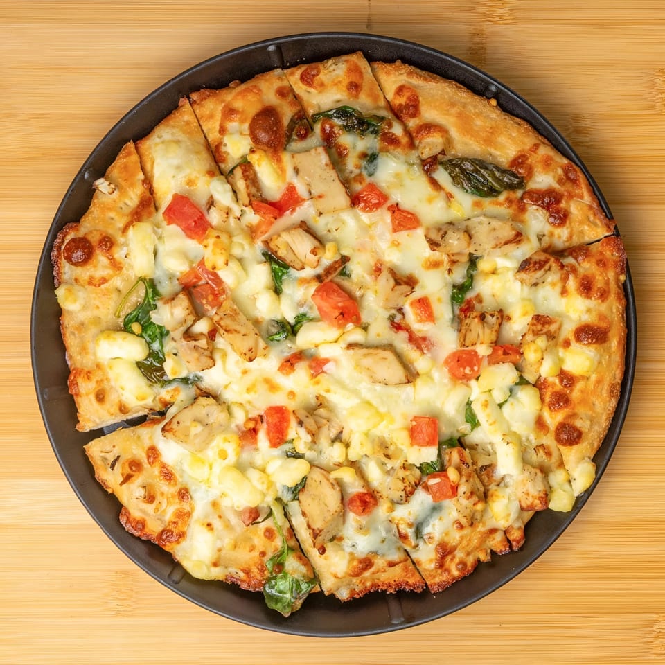 A sliced pizza with chicken, tomatoes, and cheese on a black plate, on a wooden surface.