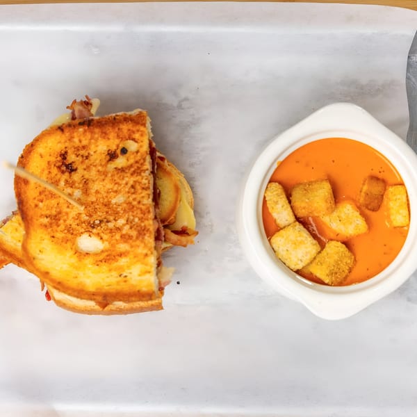 Grilled cheese sandwich with bacon and melted cheese, next to a bowl of tomato soup with croutons.