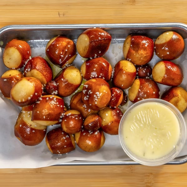 A metal tray filled with salted pretzel bites and a small cup of cheese dipping sauce.